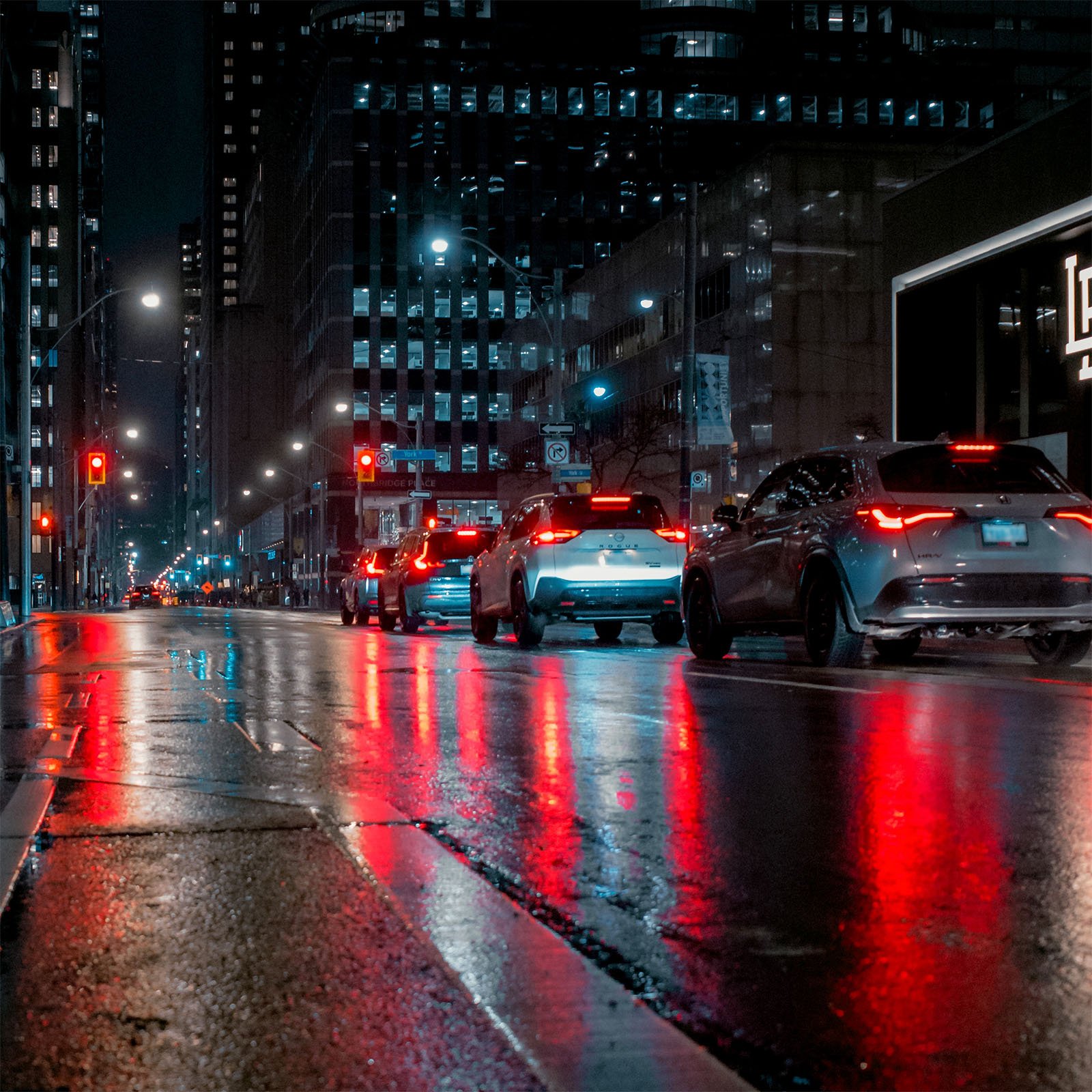 Cars wait at a traffic light on a wet city street at night, with reflections of red and white lights on the pavement. Tall buildings with illuminated windows line the street, contributing to the urban ambiance.