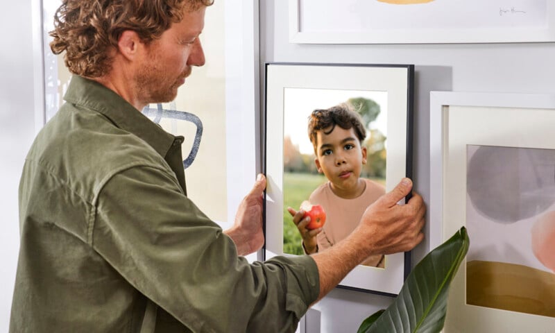 A man adjusts a framed photo of a young boy holding an apple on a gallery wall filled with various artworks inside a bright, modern room.