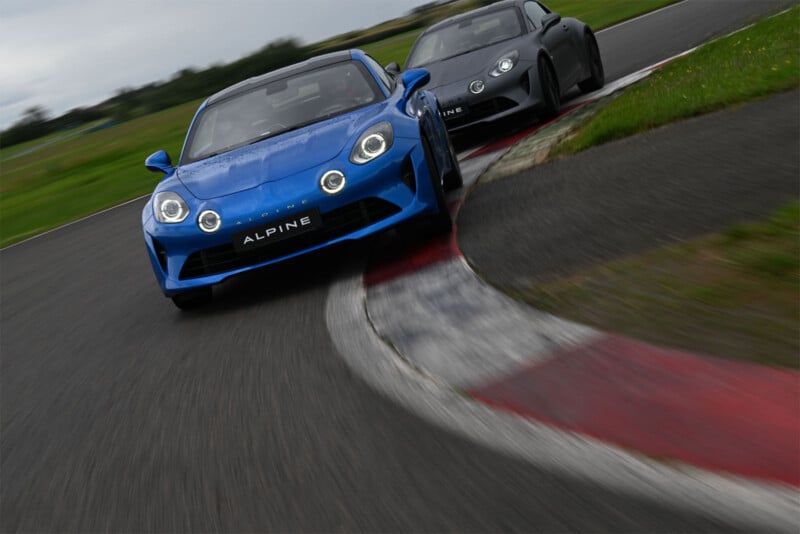 A photo of two cars racing against each other. The car in the front is blue, and the cars are going around a corner. 