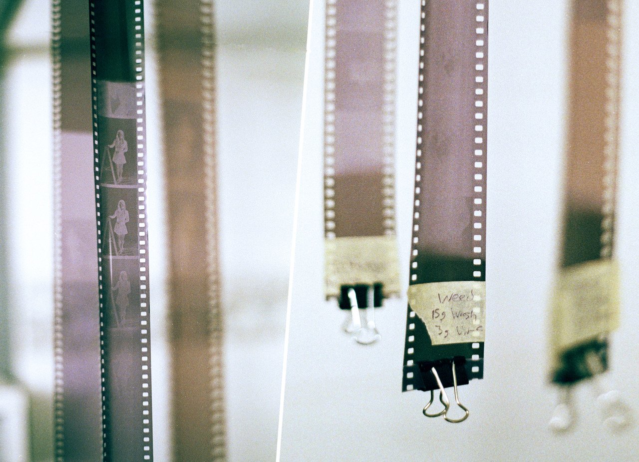 Strips of film negatives hanging from clips with handwritten labels, showcasing various frames and scenes, creating an artsy backdrop with a focus on photographic development.