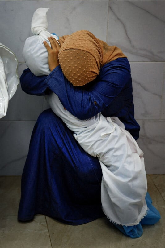 A woman dressed in blue and a yellow hijab, mourns, sitting, cradling her head in her arms while holding a deceased child wrapped in white cloth.