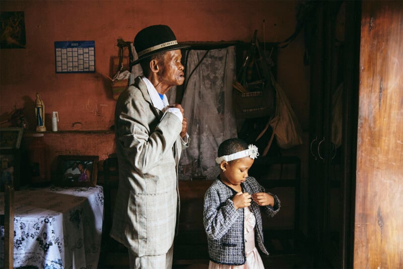 An elderly man and a young girl, both dressed in formal clothing, stand in a rustic room, gazing down as they adjust their attire, with sunlight filtering in through a window.