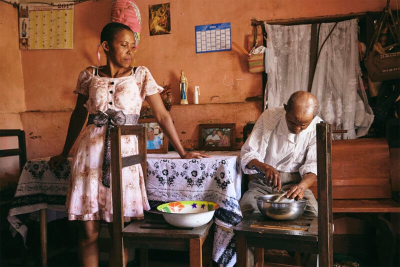 A woman in a floral dress and a man in a white shirt prepare food in a rustic kitchen, filled with homey decor and cooking utensils.