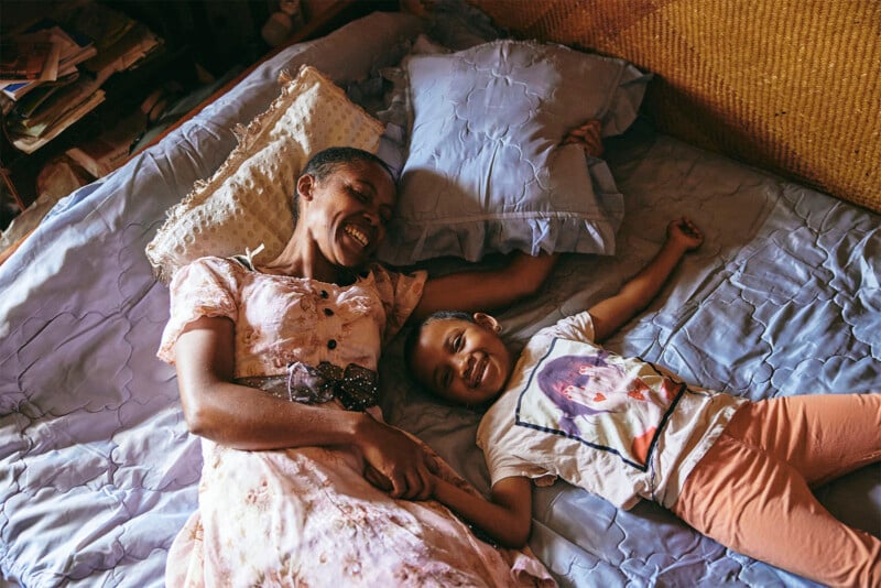 A mother and daughter laugh joyfully while lying down together on a bed in a warmly lit room filled with books and pillows.