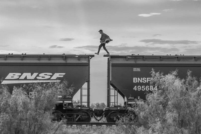 A person balances atop a moving freight train, carefully walking along the narrow edge between two train cars under a cloudy sky.