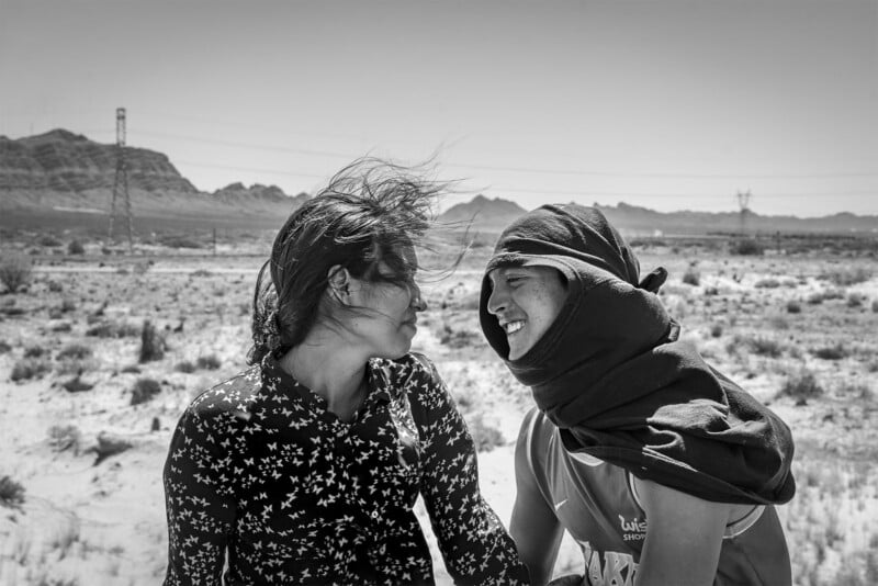 Two people smiling and laughing together in a windy desert landscape, with one woman's hair blowing across her face. power lines and sparse vegetation are visible in the background.