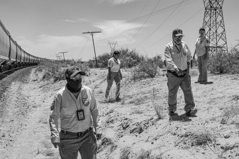 Three immigration officers in uniforms, one in the foreground wearing a face mask, stand by a railway track in a desert area with power lines and sparse vegetation.