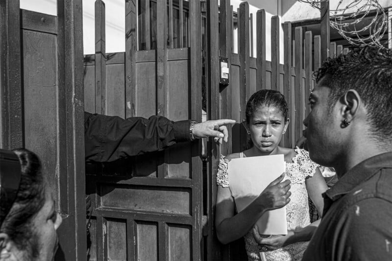 A young girl, visibly distressed, stands at a gate receiving a document from a hand extending through the bars, as a man looks on seriously. this black and white photo captures a tense, emotional moment.