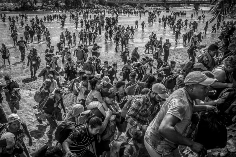 Black and white image of a crowded river crossing where numerous people, carrying belongings, wade through water under a bridge, depicting a scene of migration or displacement.