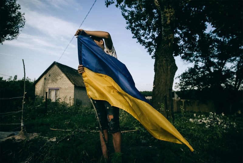 A person obscured by a large ukrainian flag, standing in a rural area with trees and a house in the background during twilight.