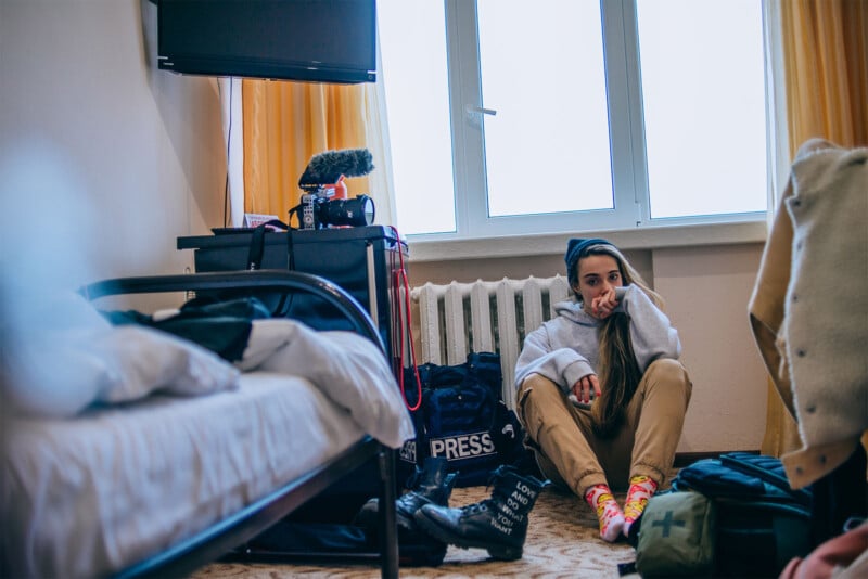 A photojournalist sits on the floor of a modestly furnished hotel room, looking thoughtful. nearby is a camera on a tripod and gear labeled "press." clothes and a pair of boots add to the cluttered space.