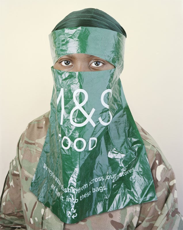 A person wearing a camouflage jacket is partially covered by a green m&s plastic bag fashioned as a veil, with only their eyes visible, against a neutral background.