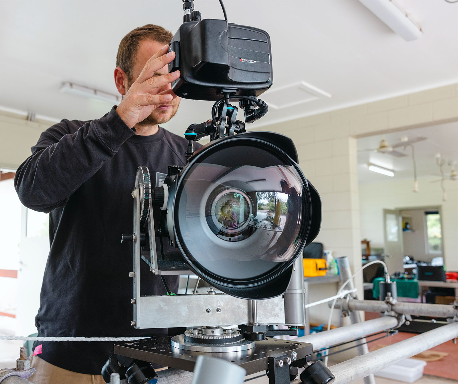 A man adjusts a large lens on a professional camera mounted on a tripod in a well-lit room, focusing with his hand partially covering his face.