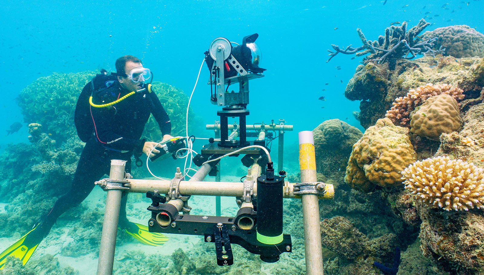 A scuba diver operates scientific underwater equipment near vibrant coral reefs in clear blue water.