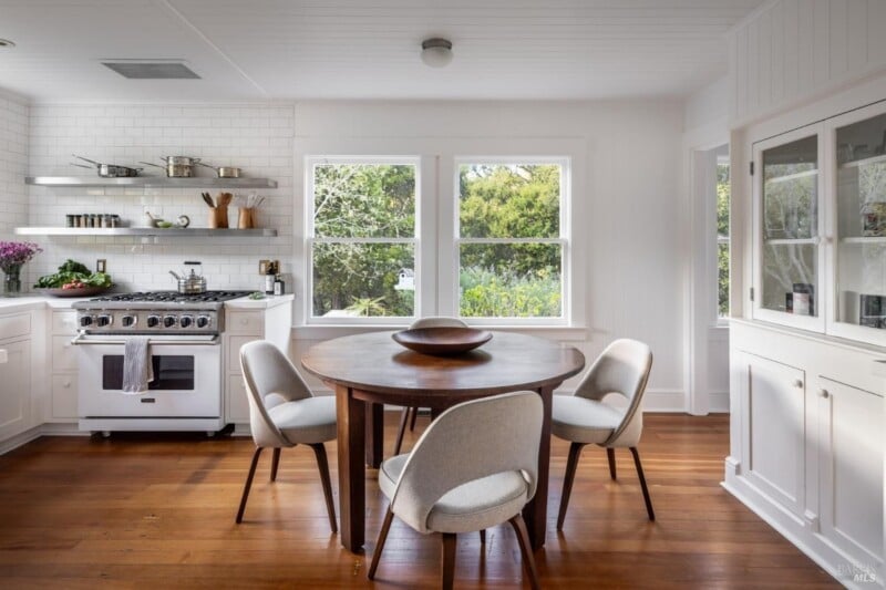 A bright, modern kitchen with white cabinetry and subway tile backsplash, featuring a stainless-steel stove, wooden dining table with grey chairs, and large windows with a greenery view.
