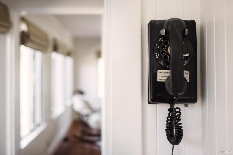 An old-fashioned black rotary dial telephone hangs on a white wall in a narrow hallway with windows, casting soft light across the scene.