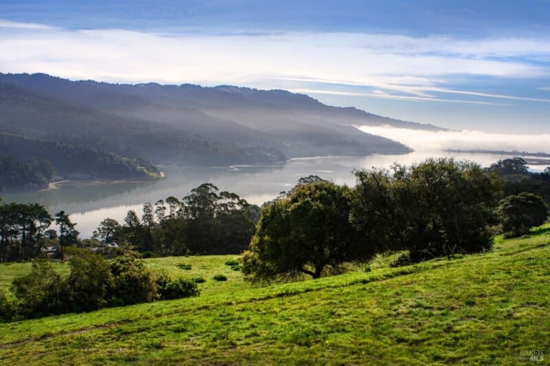 A serene landscape featuring a river winding through lush green hills under a blue sky, with patches of morning mist clinging to the valley.