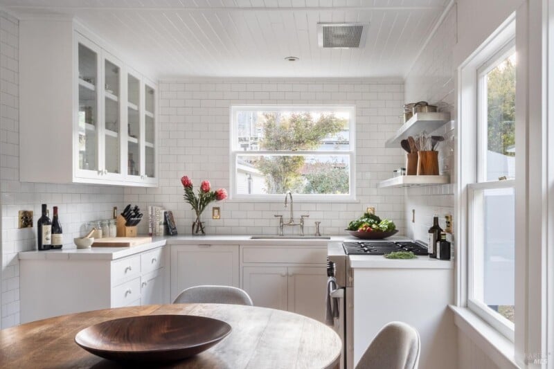 A bright, modern kitchen with white cabinetry, subway tiles, and a round wooden dining table. large windows allow natural light to illuminate the space.
