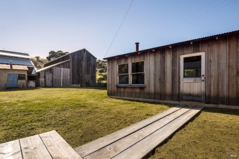 Rustic wooden buildings on a green grassy field under a clear blue sky, connected by a wooden plank walkway, portray a serene countryside setting.