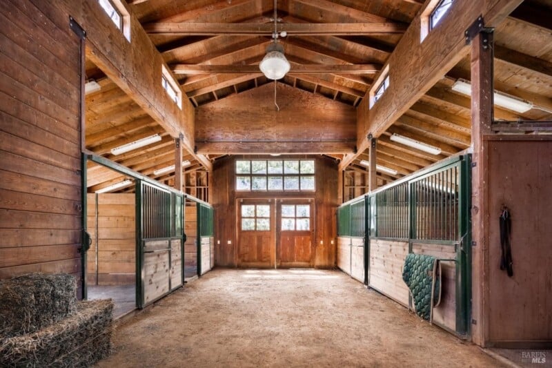 Interior of a clean, well-lit stable with wooden walls, a high ceiling with a fan, and several stalls. a hay bale and saddle pads are also visible.
