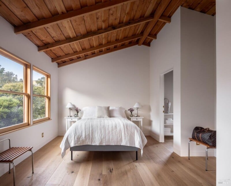 A serene bedroom with a wooden beamed ceiling, beige walls, large windows, and hardwood floors. the room features a double bed with white bedding, two nightstands, and a wooden bench.