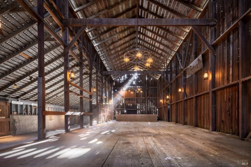 Interior of a spacious, rustic barn with a wooden structure, featuring large beams and panels, with sunlight streaming through gaps in the roof, casting patterns on the floor.