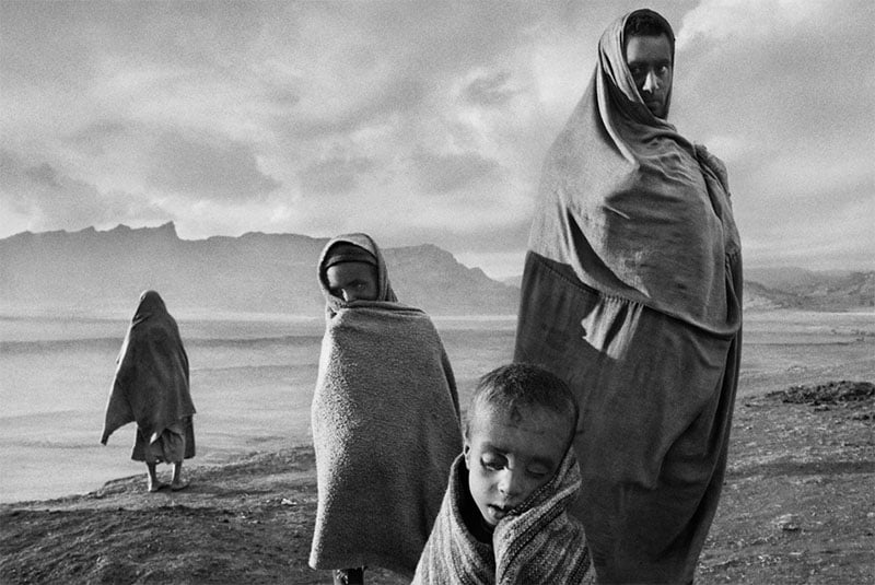 A black and white photo depicting four figures draped in cloaks on a barren landscape. their expressions are somber, and the cloudy sky adds a dramatic effect to the scene.