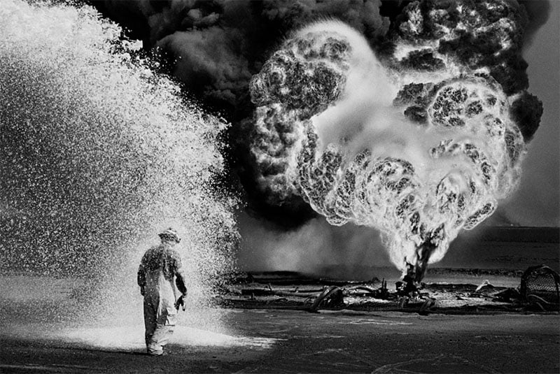 A firefighter in protective gear walks past a foreground of spraying water with a large, fiery explosion in the background, creating thick smoke that forms a dramatic contrast.