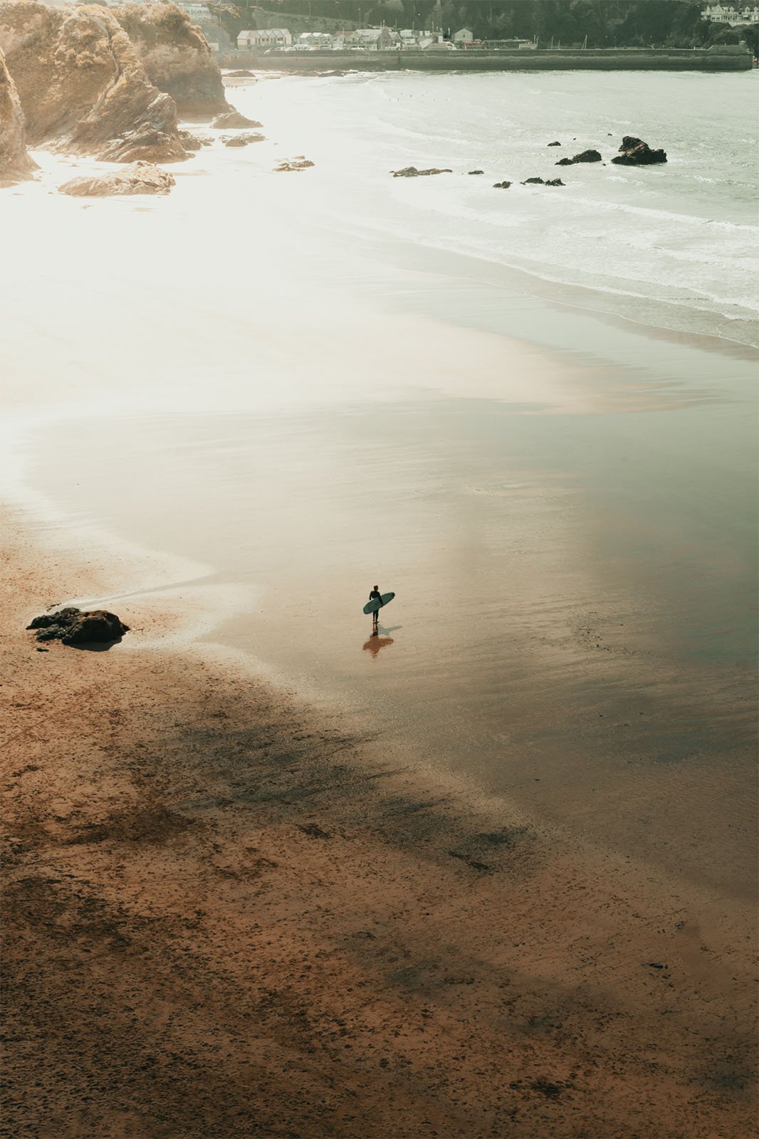 A lone surfer walks along a sunlit beach towards the water, with large rocks scattered along the shoreline, under a soft golden haze.