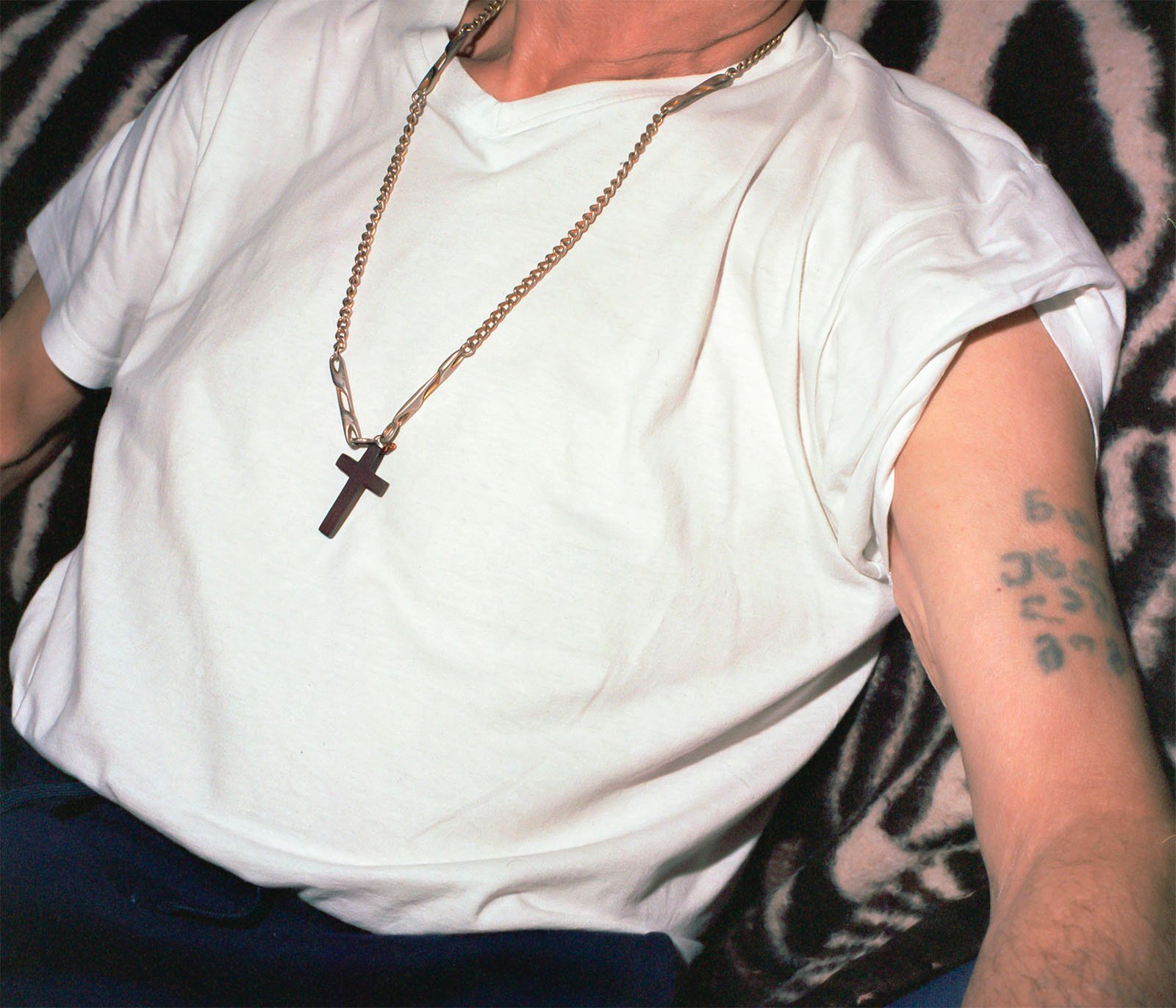 A close-up photo of a person wearing a plain white t-shirt with a golden chain and cross pendant. there's a visible tattoo with numbers on the forearm. the background features a leopard print fabric.