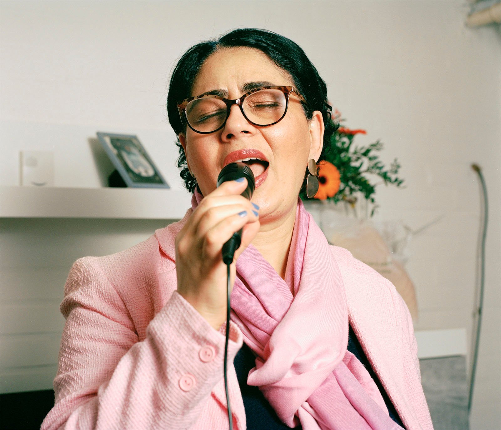 A woman wearing glasses and a pink blazer sings passionately into a microphone, eyes closed, in a room with a white wall and subtle decor.