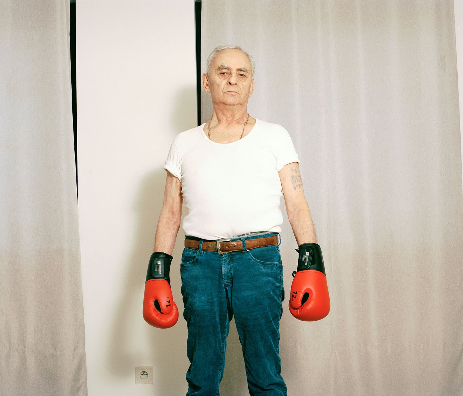 An elderly man with gray hair, wearing a white t-shirt, blue jeans, and a belt, stands with red boxing gloves on, posing confidently in front of a neutral curtain backdrop.