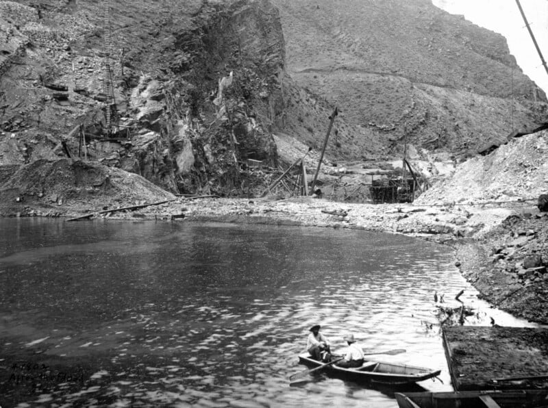 Black and white historical photo showing two people in a rowboat on a small body of water surrounded by rocky cliffs and industrial structures.