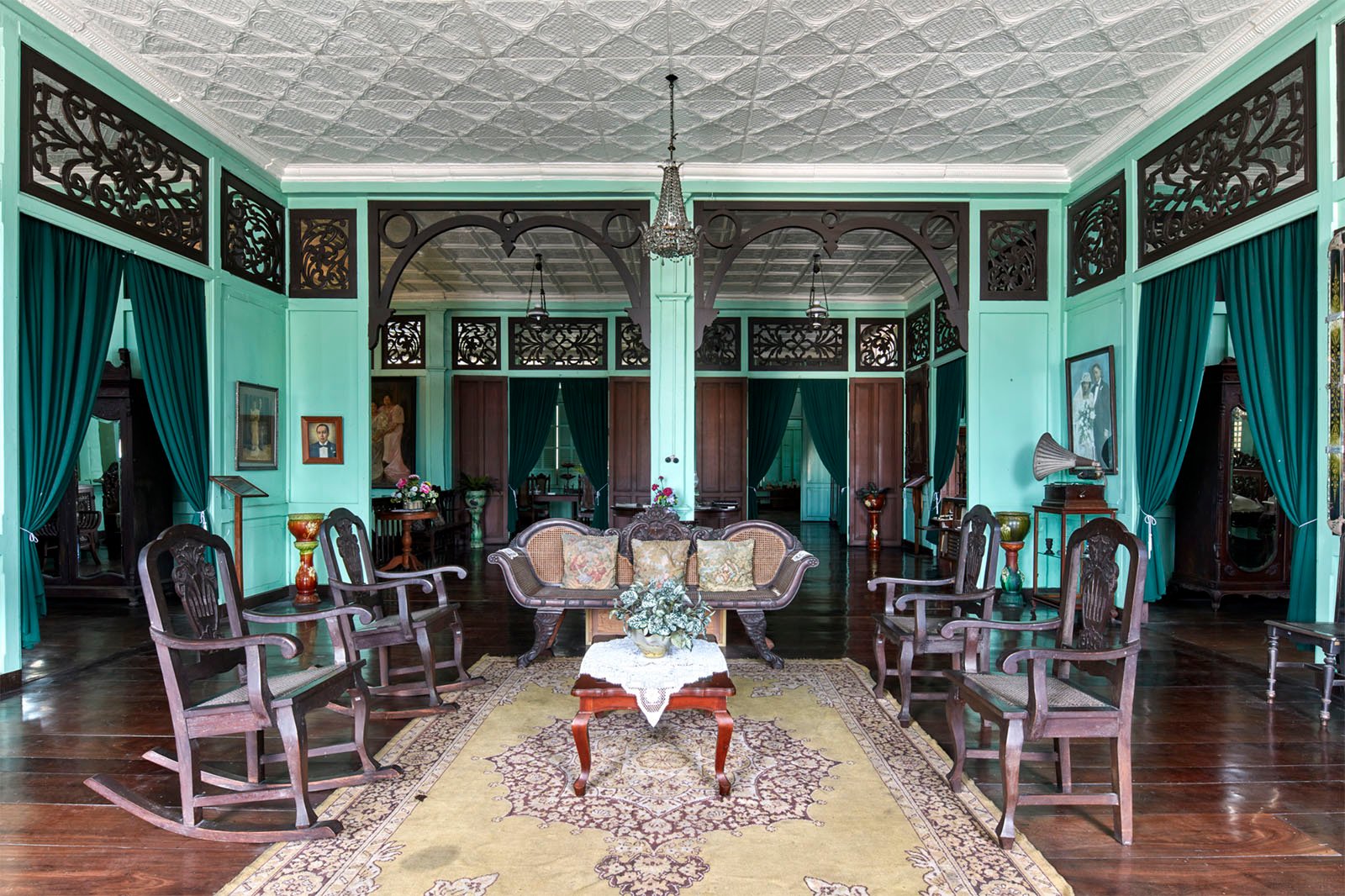 A traditional, elegant interior of a colonial-style room with wooden furniture, patterned ceiling, and large draped windows, featuring dark wooden armchairs and decorative wall panels.