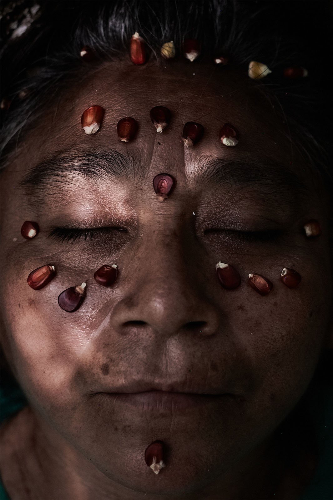 Close-up portrait of a person's face covered with various grains of corn arranged in patterns, with closed eyes. the image is dark and moody, focusing on the unique corn decoration on their skin.