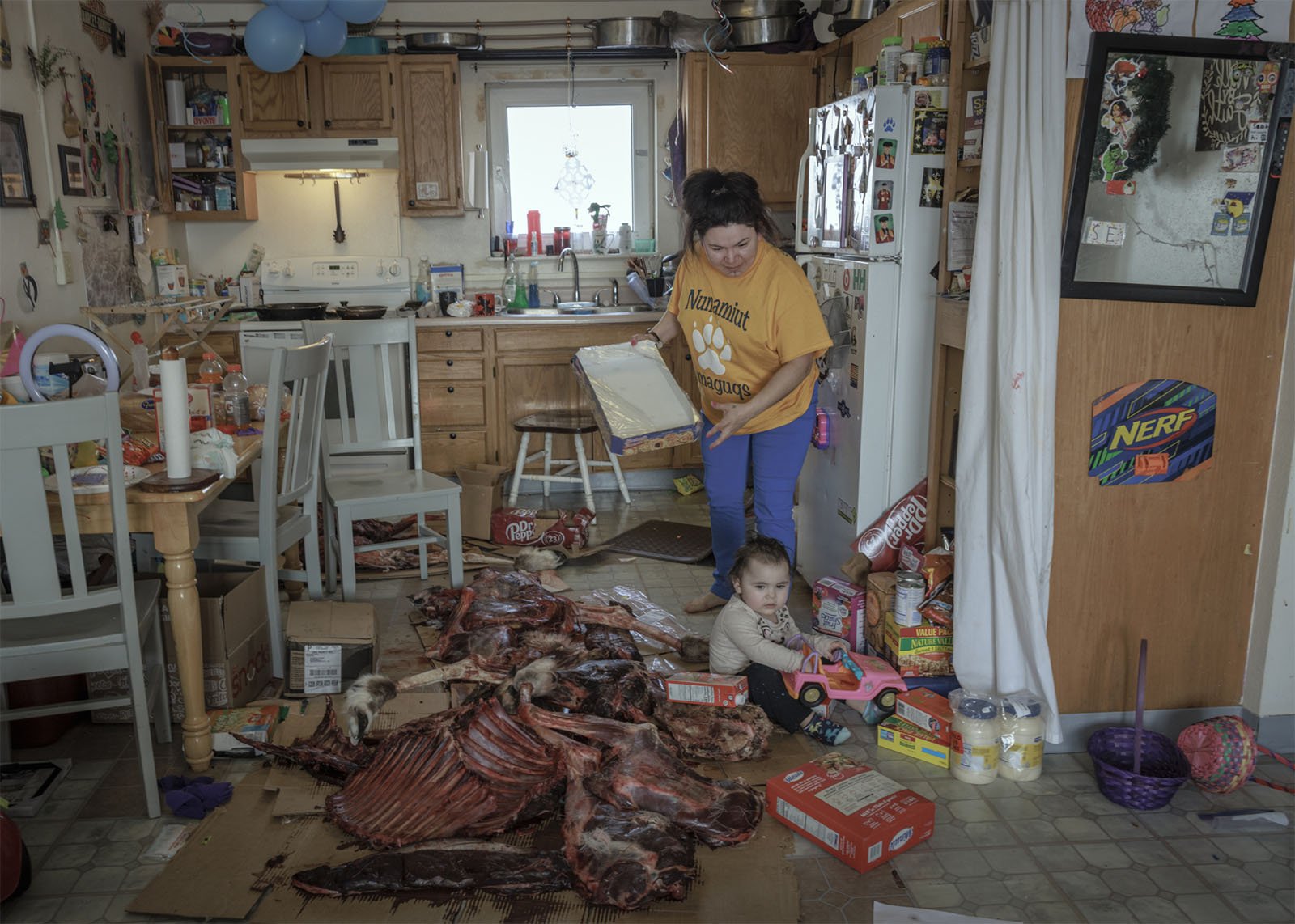 A woman and a toddler in a cluttered kitchen, where the woman is reading a recipe and the child is sitting among scattered groceries and a large pile of raw meat on cardboard.
