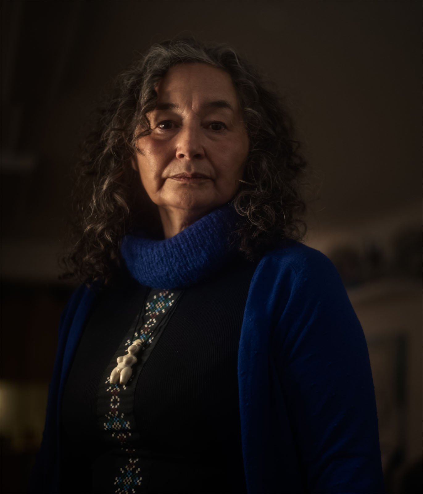 Portrait of an elderly woman with curly gray hair, wearing a blue scarf and a black shirt, looking solemnly at the camera in a dimly lit room.