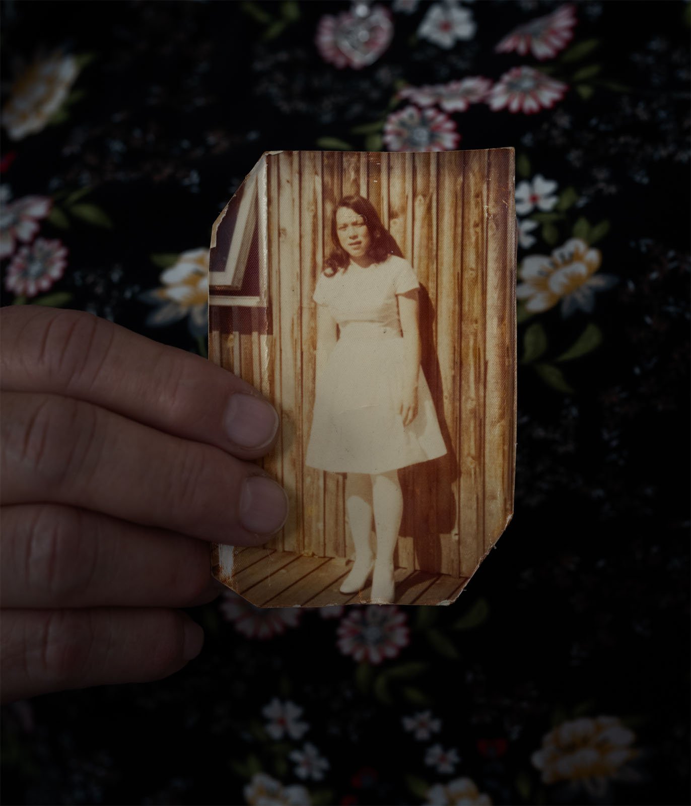 A hand holds a worn, vintage photograph of a woman standing in front of a wooden backdrop, against a dark floral patterned background.