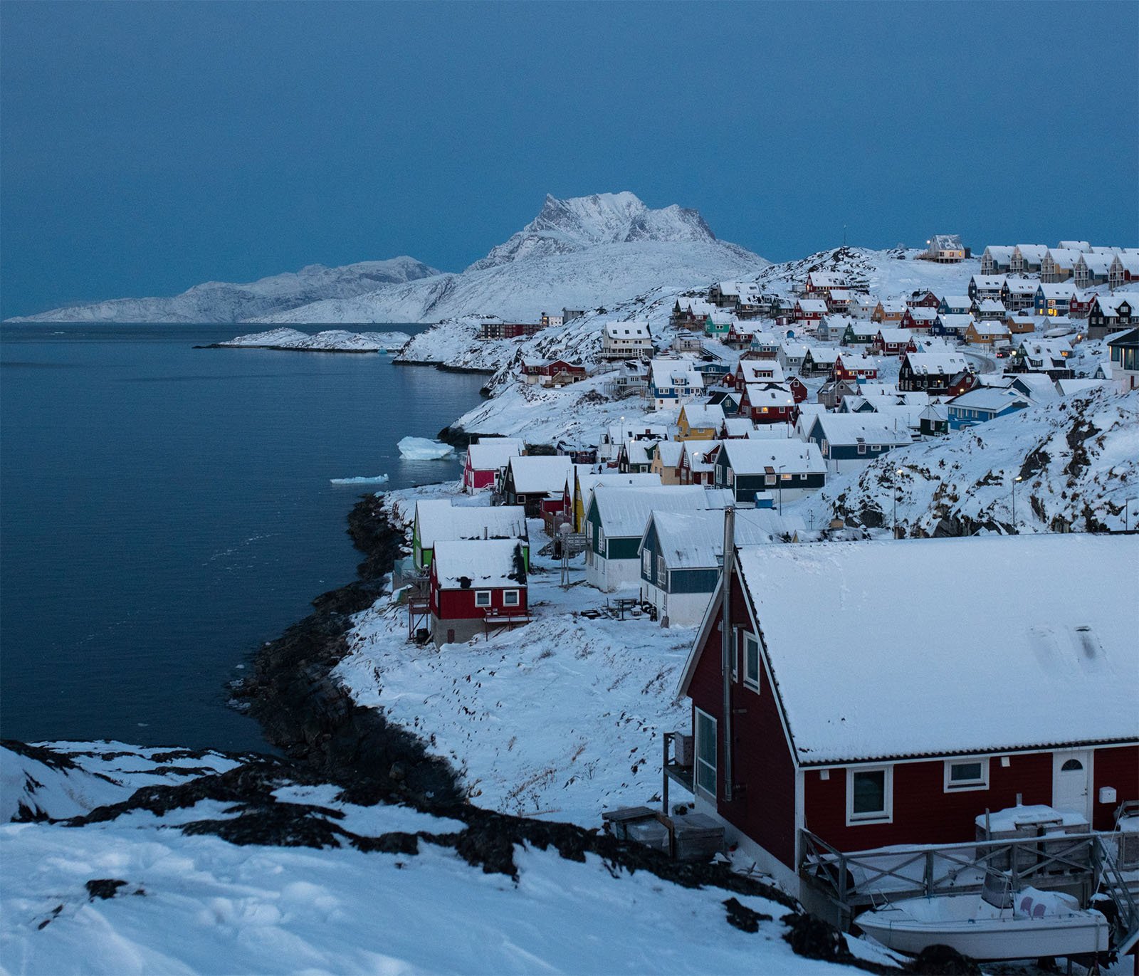 A snowy coastal village with colorful houses overlooking a serene bay, under a dusky blue sky, with rugged mountains in the background.