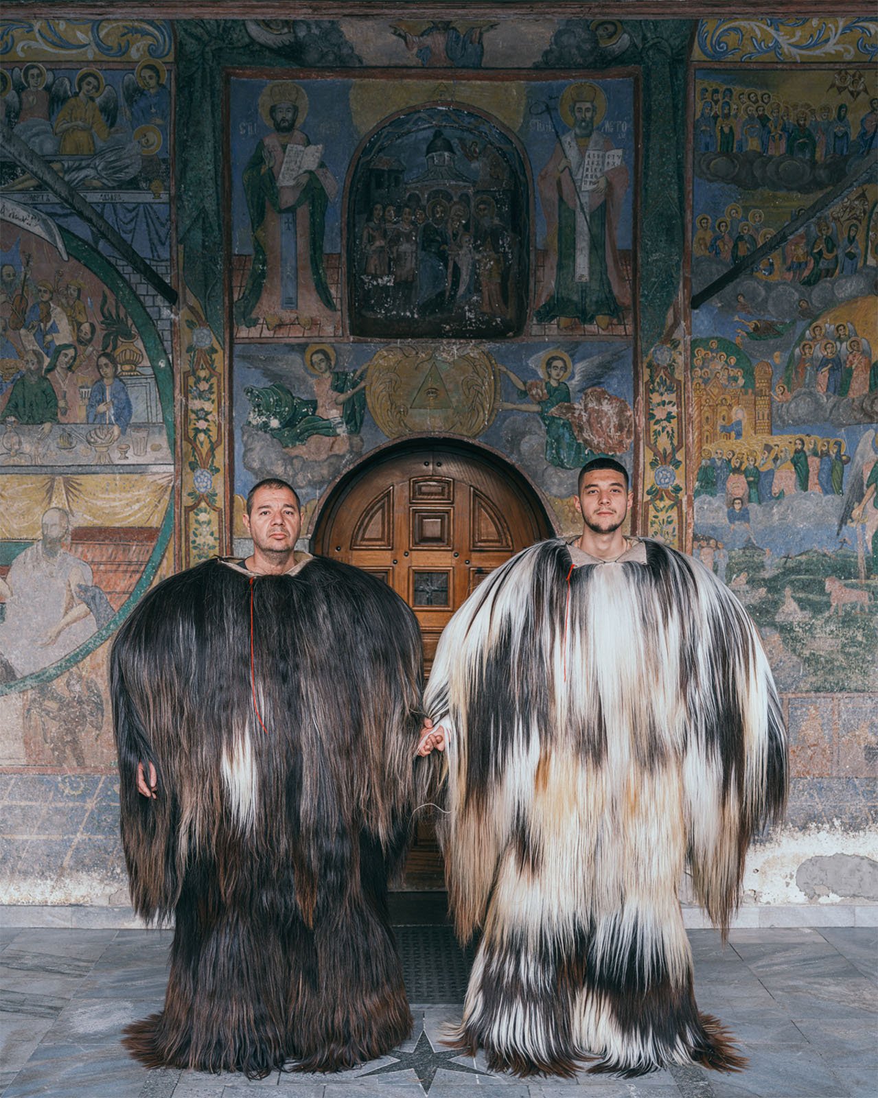Two men stand in front of an ornately decorated church entrance, wearing traditional long, shaggy wool garments. the wall behind them features vibrant religious frescoes.
