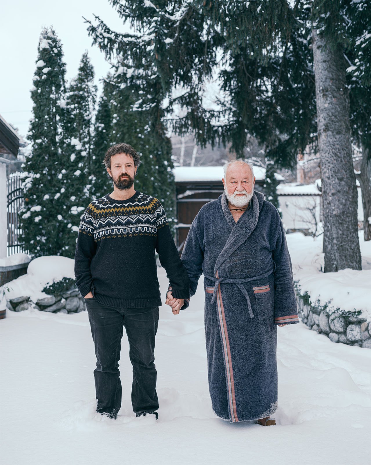 A young man in a sweater and an older man in a bathrobe standing hand in hand on a snowy path, with tall pine trees and a light snowfall around them.