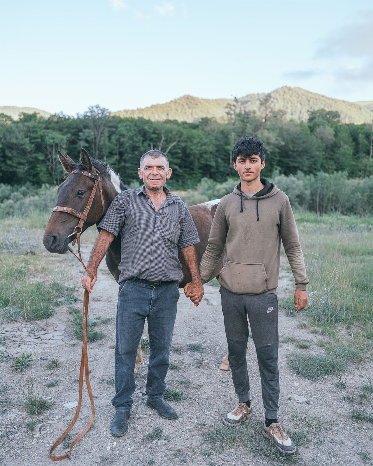 An elderly man and a young man hold hands, standing next to a brown horse in a natural setting with trees and a hill in the background. they both face the camera with subtle smiles.