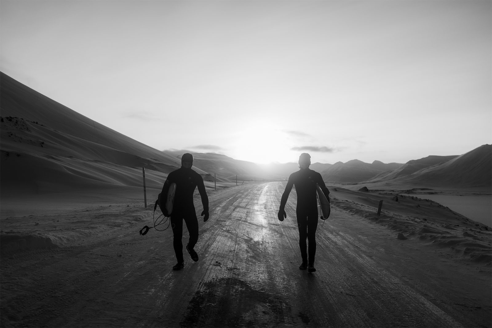 Two surfers in wetsuits walking on a snowy road towards a setting sun, with snow-covered hills on either side.