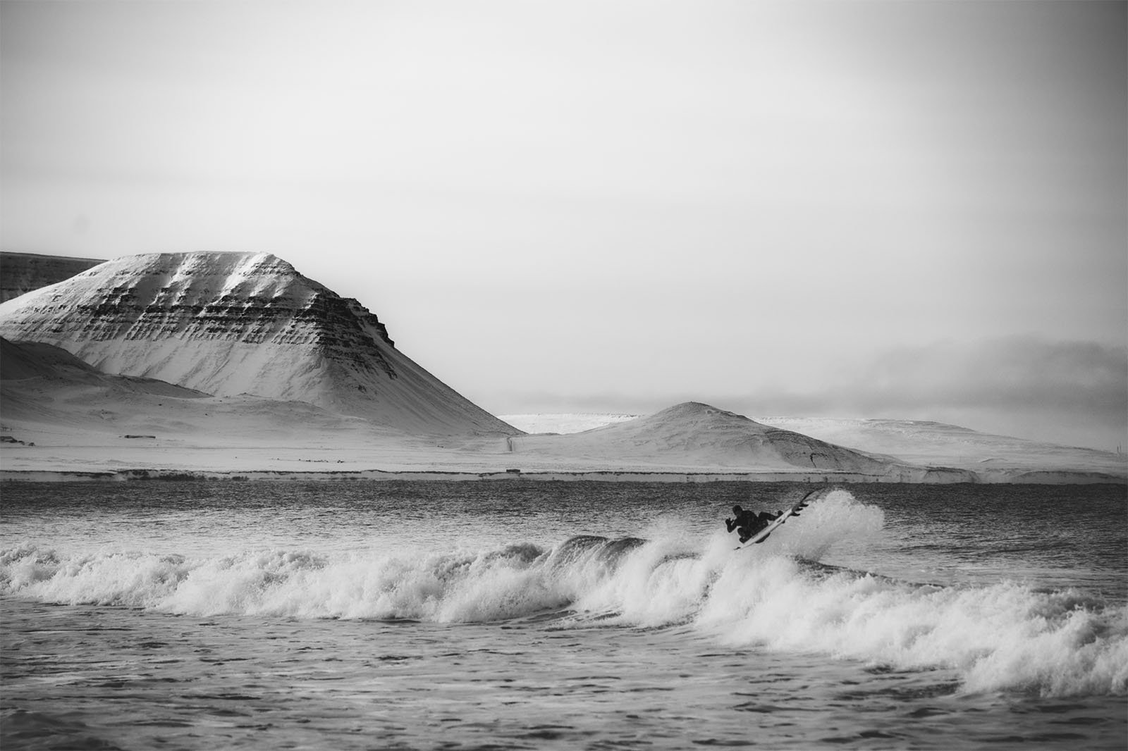 A surfer rides a wave in a cold, misty sea with snow-capped mountains in the background under an overcast sky.
