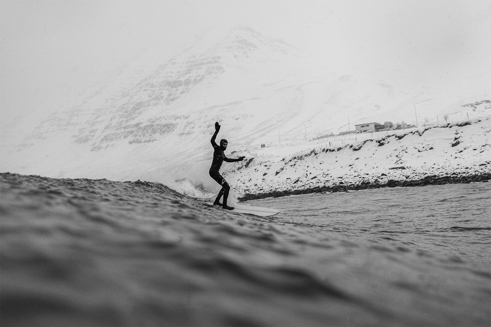 A surfer rides a wave in a wintery landscape, with snow-covered mountains in the background and shore visible. the image captures a moment of contrast between the snowy environment and the aquatic activity.