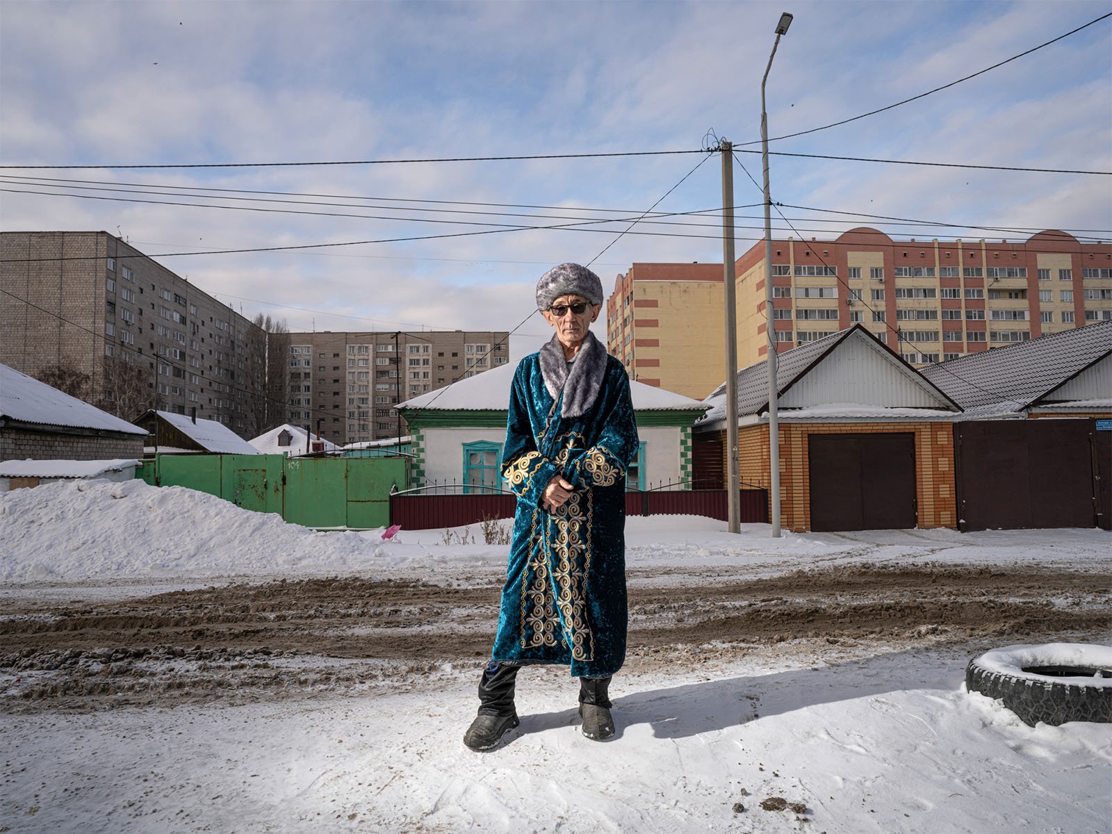 An elderly woman dressed in a traditional robe and hat stands confidently on a snowy street, with colorful apartment buildings in the background under a partly cloudy sky.