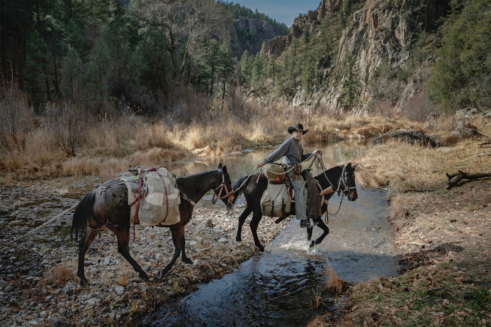 A cowboy on horseback leading another horse across a shallow river in a rugged canyon landscape with wooded slopes and rocky terrain.