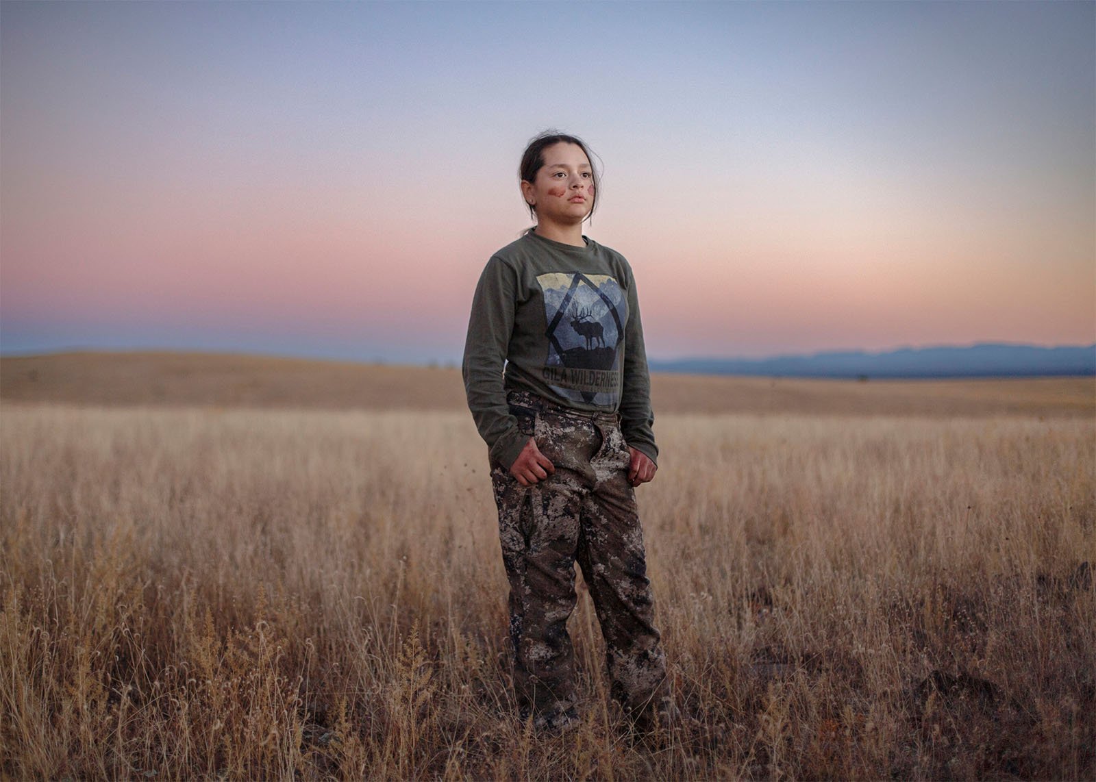A young woman stands in a field at twilight, wearing camouflage pants and a green shirt with a skull design, looking confidently at the camera with a serene sky behind her.