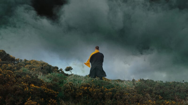 A man stands in a field with dramatic clouds behind him.