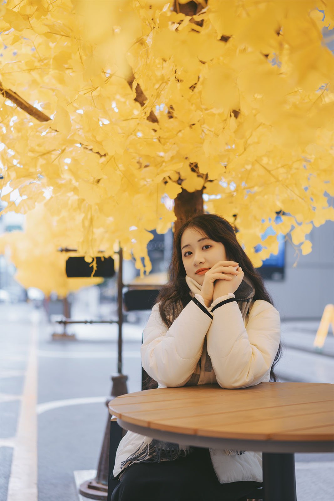 A young woman sits at an outdoor table under a canopy of bright yellow autumn leaves, her elbows on the table and hands under her chin, looking thoughtful.